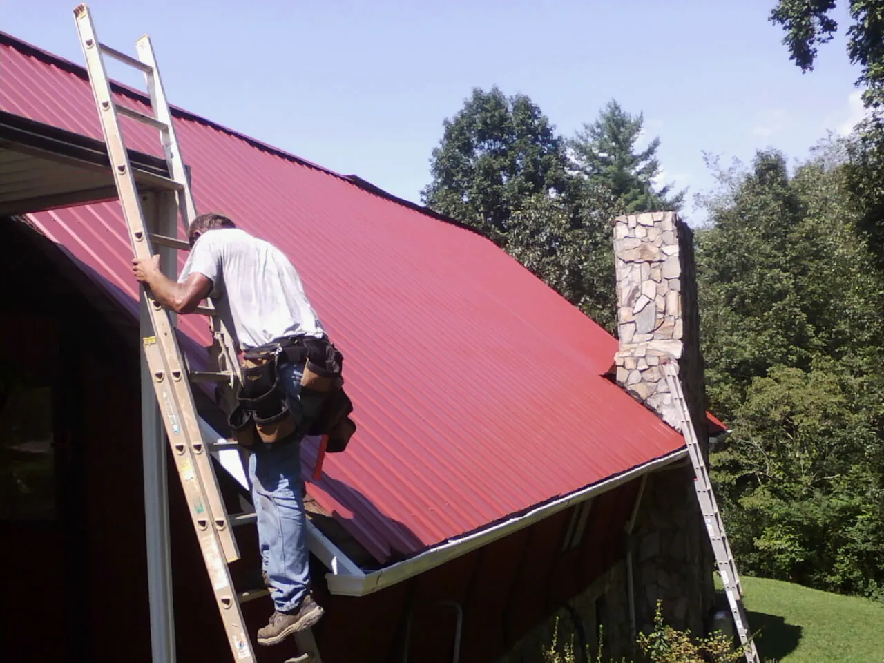 Roofer on a ladder beside a red metal roof and stone chimney — Soffit & Fascia Repair by Roofing Brothers in Springfield Gardens